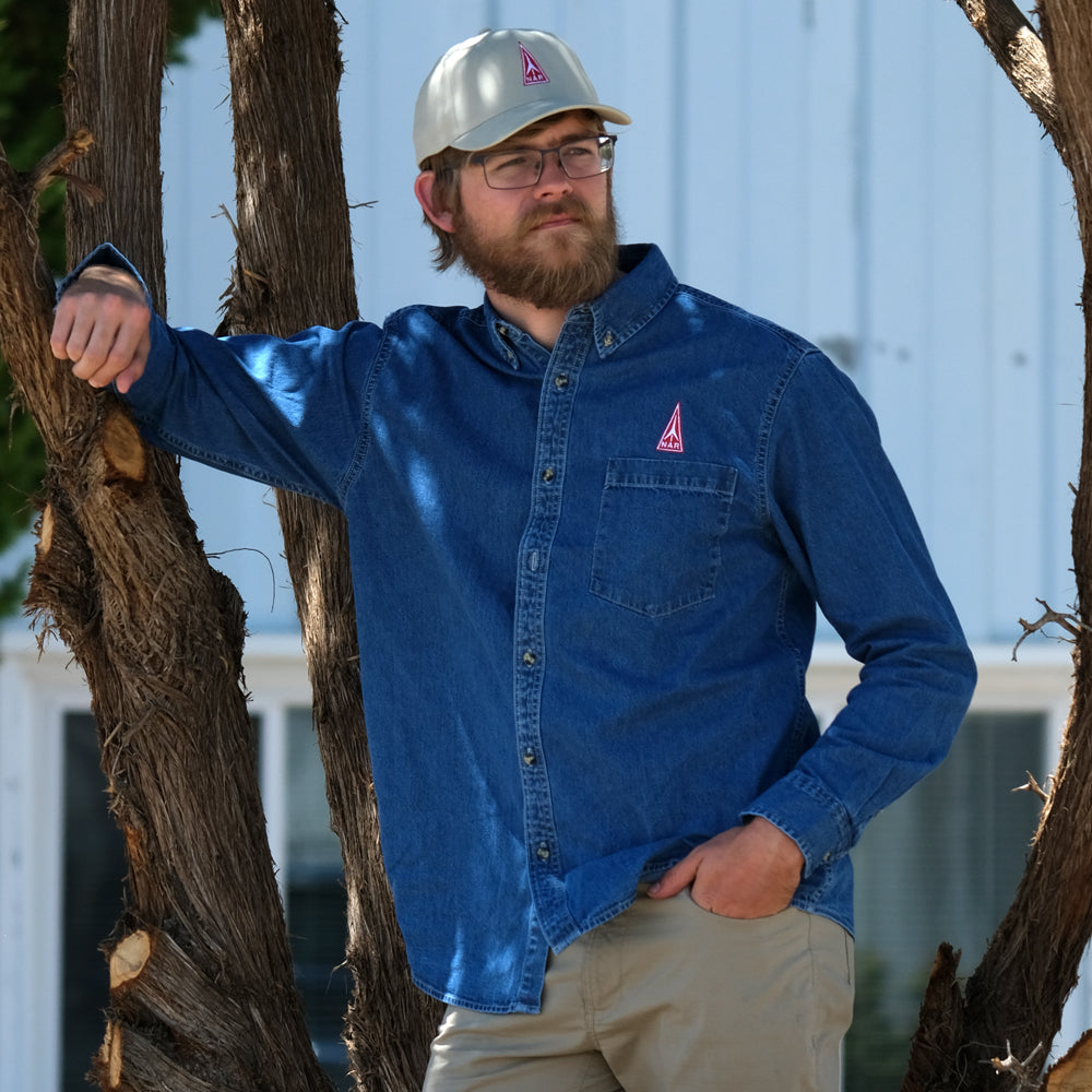 Man wearing a National Association of Rocketry Denim Shirt and NAR cap leaning against a tree.
